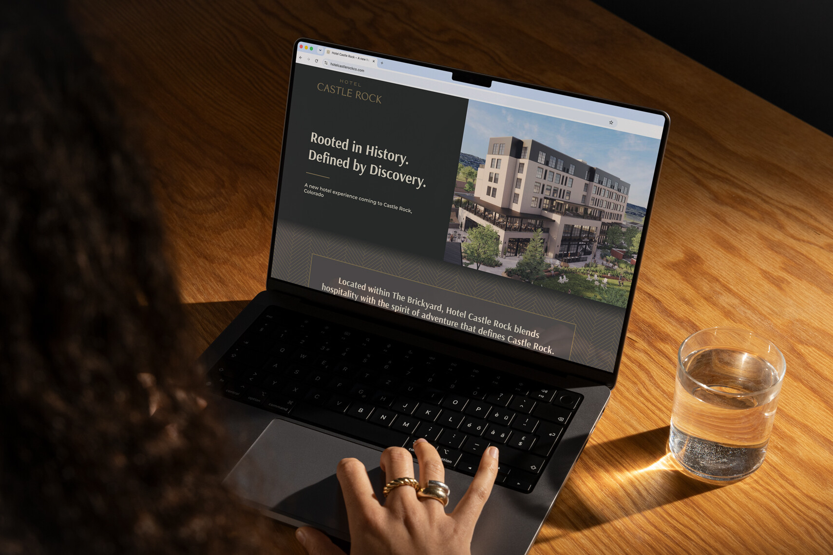 Woman using her laptop to view the Hotel Castle Rock website at a desk with a glass of water